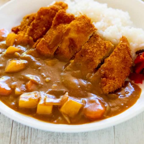 A bowl of Japanese katsu curry, featuring crispy breaded chicken cutlets served over white rice with a side of rich curry sauce containing diced carrots, garnished with green onions.